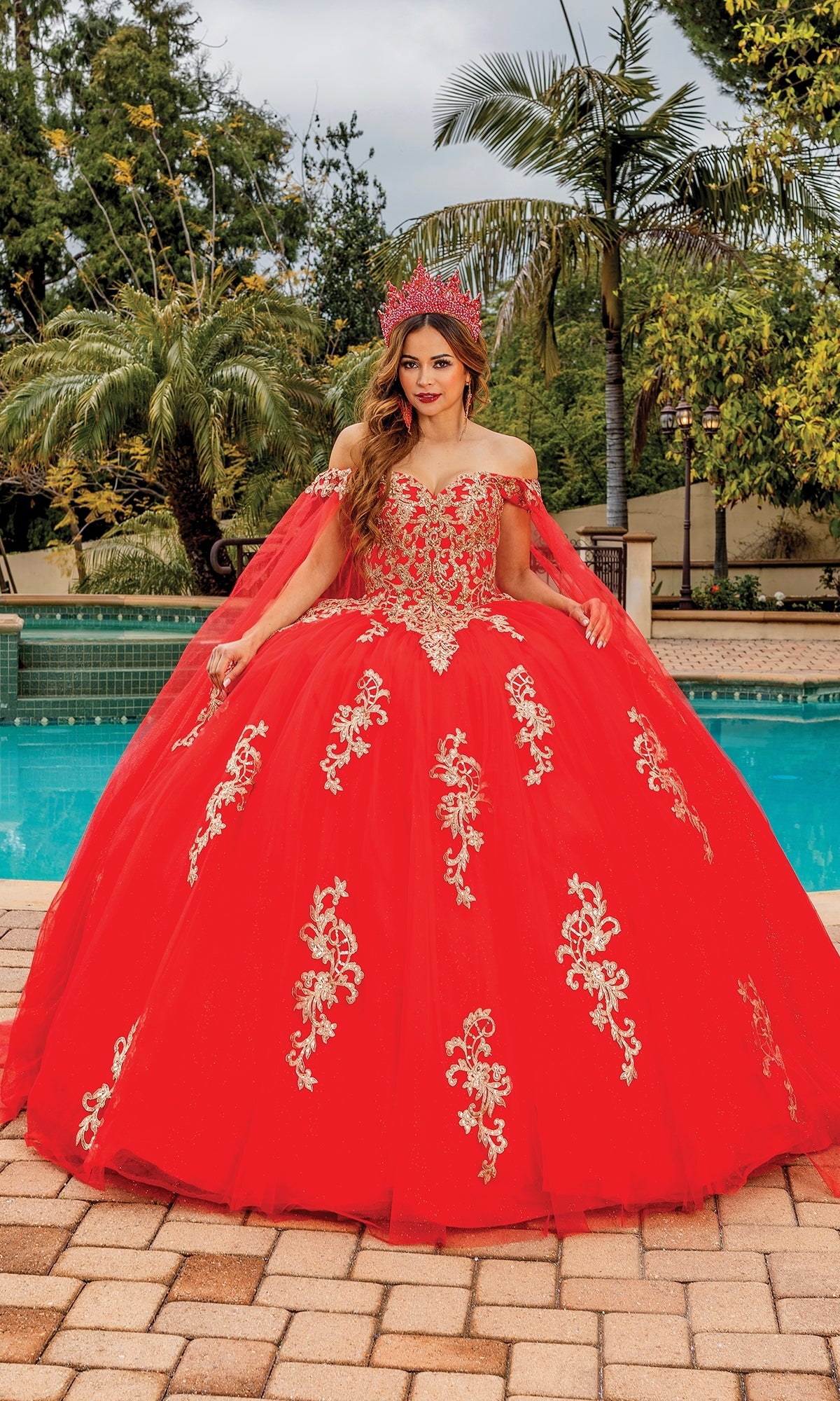 A woman stands by a pool among palm trees, wearing the Quinceanera Dress 1740 By Dancing Queen—a bright red off-the-shoulder gown with gold embroidery and draped cape sleeves.