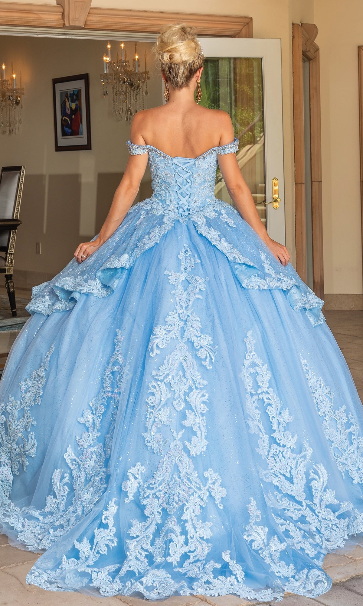 A woman stands indoors facing away from the camera, wearing Quinceanera Dress 1741 by Dancing Queen—a light blue, ornate A-line ball gown with lace detailing and off-the-shoulder straps.