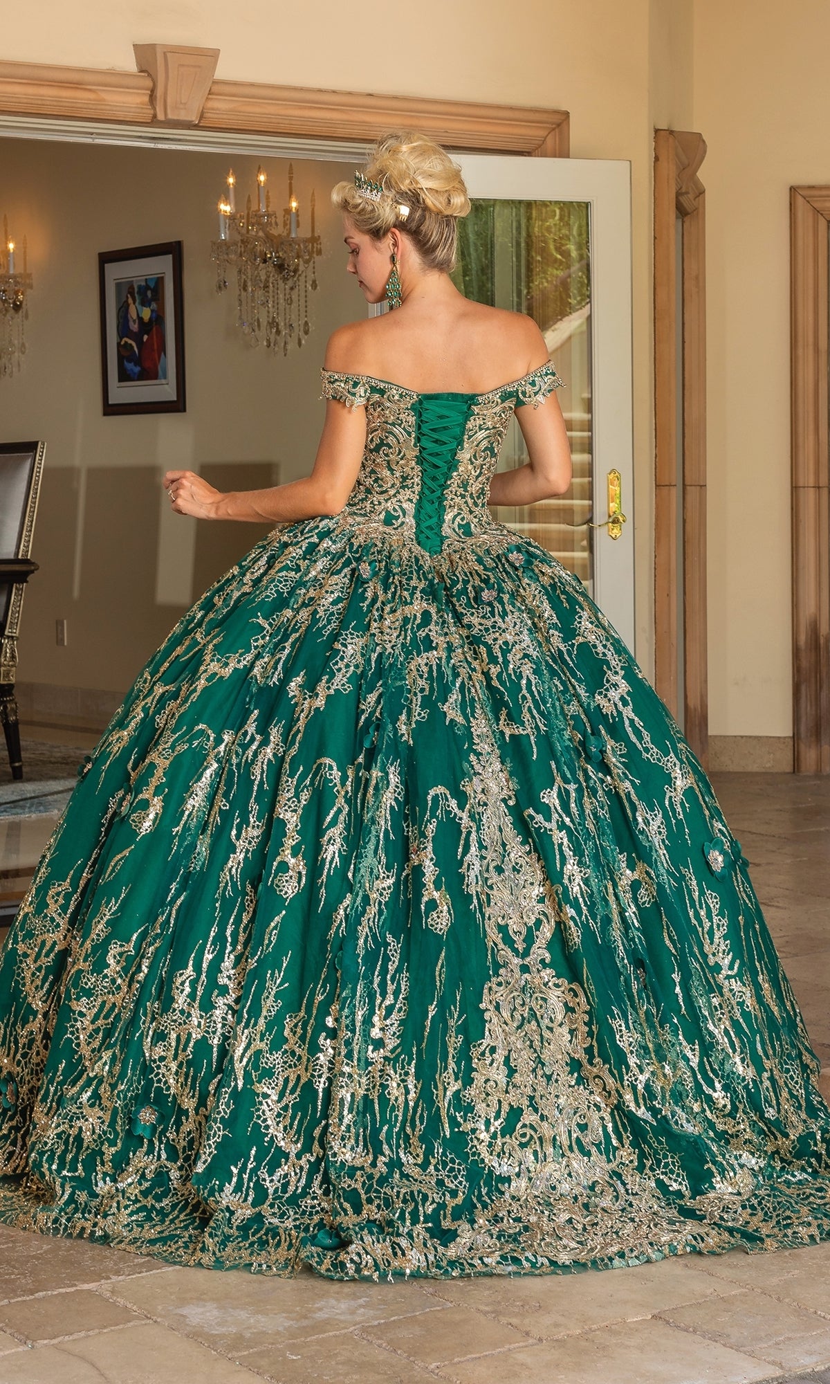 A woman stands indoors wearing the Quinceanera Dress 1742 By Dancing Queen, an emerald green lace-embroidered ball gown with off-the-shoulder straps, gold detailing, and a flowing full skirt as she faces away from the camera.