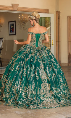 A woman stands indoors wearing the Quinceanera Dress 1742 By Dancing Queen, an emerald green lace-embroidered ball gown with off-the-shoulder straps, gold detailing, and a flowing full skirt as she faces away from the camera.
