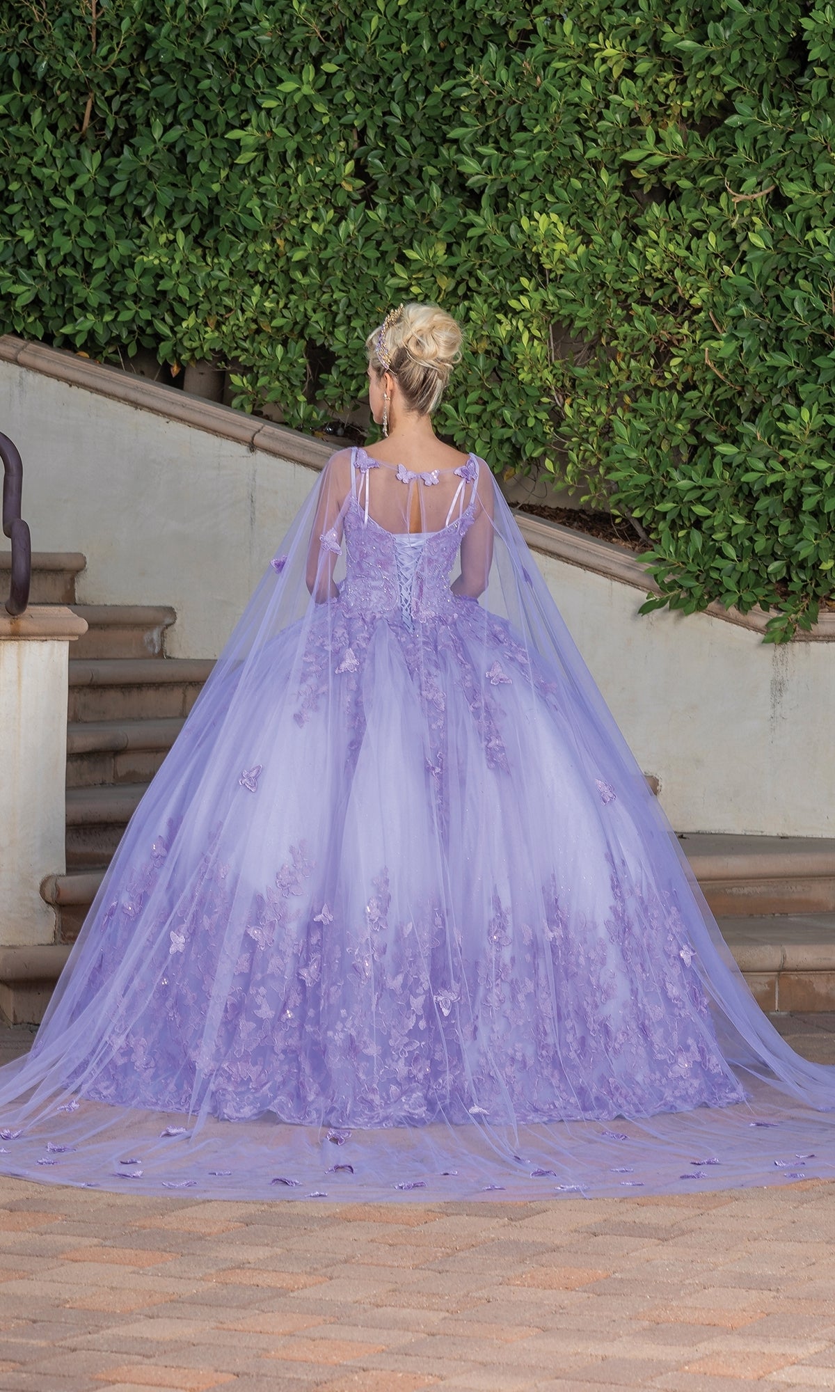 A person stands outdoors in front of steps, their back to the camera, wearing the Quinceanera Dress 1746 by Dancing Queen—a floral a-line ball gown with a long sheer cape—framed by lush green bushes.