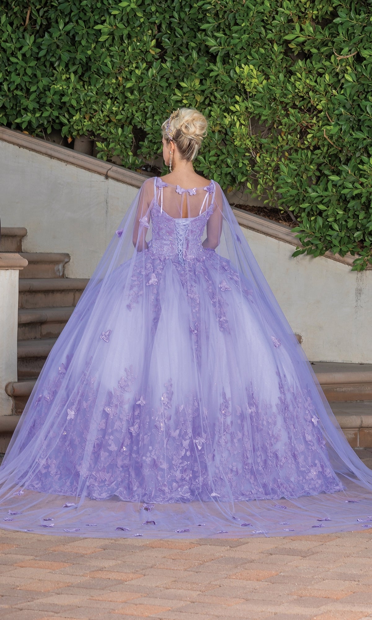 A person in the Quinceanera Dress 1746 By Dancing Queen, featuring an a-line silhouette, sweetheart neckline, floral details, and a long cape, stands on a stone walkway with greenery in the background, facing away from the camera.