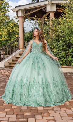 A woman stands on a brick walkway in front of a gazebo, wearing the Quinceanera Dress 1746 by Dancing Queen—a light green, A-line ball gown with floral details, a full skirt, and cape—surrounded by lush greenery.