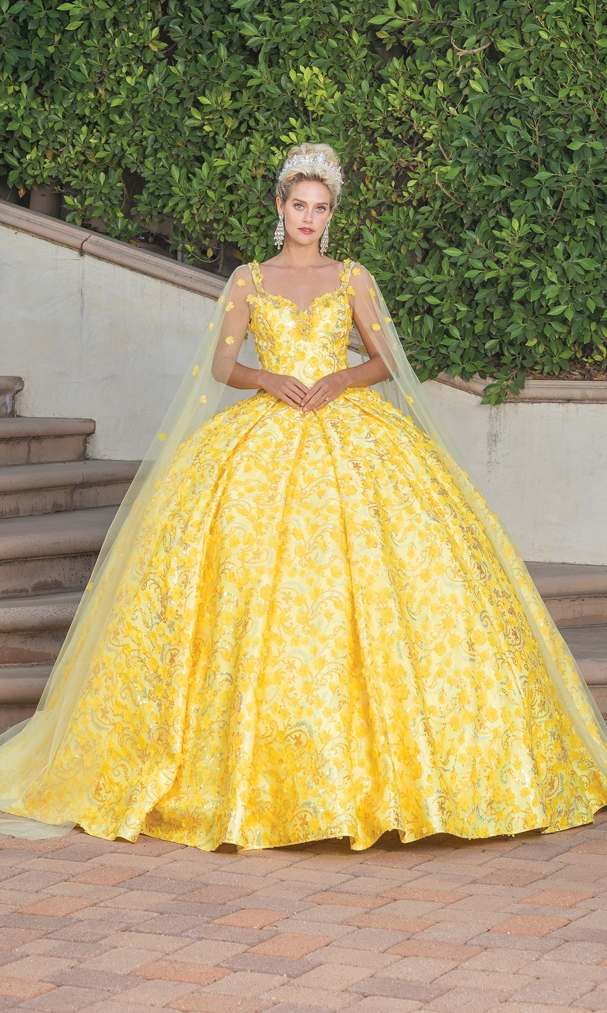 A woman stands outdoors on a stone patio in the Quinceanera Dress 1750 By Dancing Queen, a yellow ball gown with intricate patterns, sweetheart neckline, draped cape sleeves, and a tiara, surrounded by lush greenery.