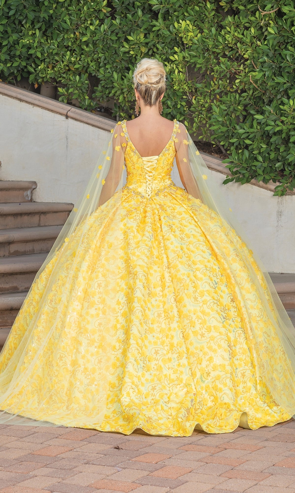 A woman in the Quinceanera Dress 1750 By Dancing Queen, featuring a yellow ball gown with a voluminous skirt, sweetheart neckline, and draped cape sleeves, stands outdoors near steps and lush greenery, facing away from the camera.