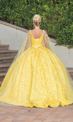 A woman in the Quinceanera Dress 1750 By Dancing Queen, featuring a yellow ball gown with a voluminous skirt, sweetheart neckline, and draped cape sleeves, stands outdoors near steps and lush greenery, facing away from the camera.