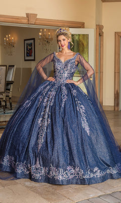 A woman wearing the Quinceanera Dress 1751 by Dancing Queen, a navy blue ball gown with silver and purple lace embroidery and sheer draped sleeves, stands indoors near an open door, accessorized with a tiara.
