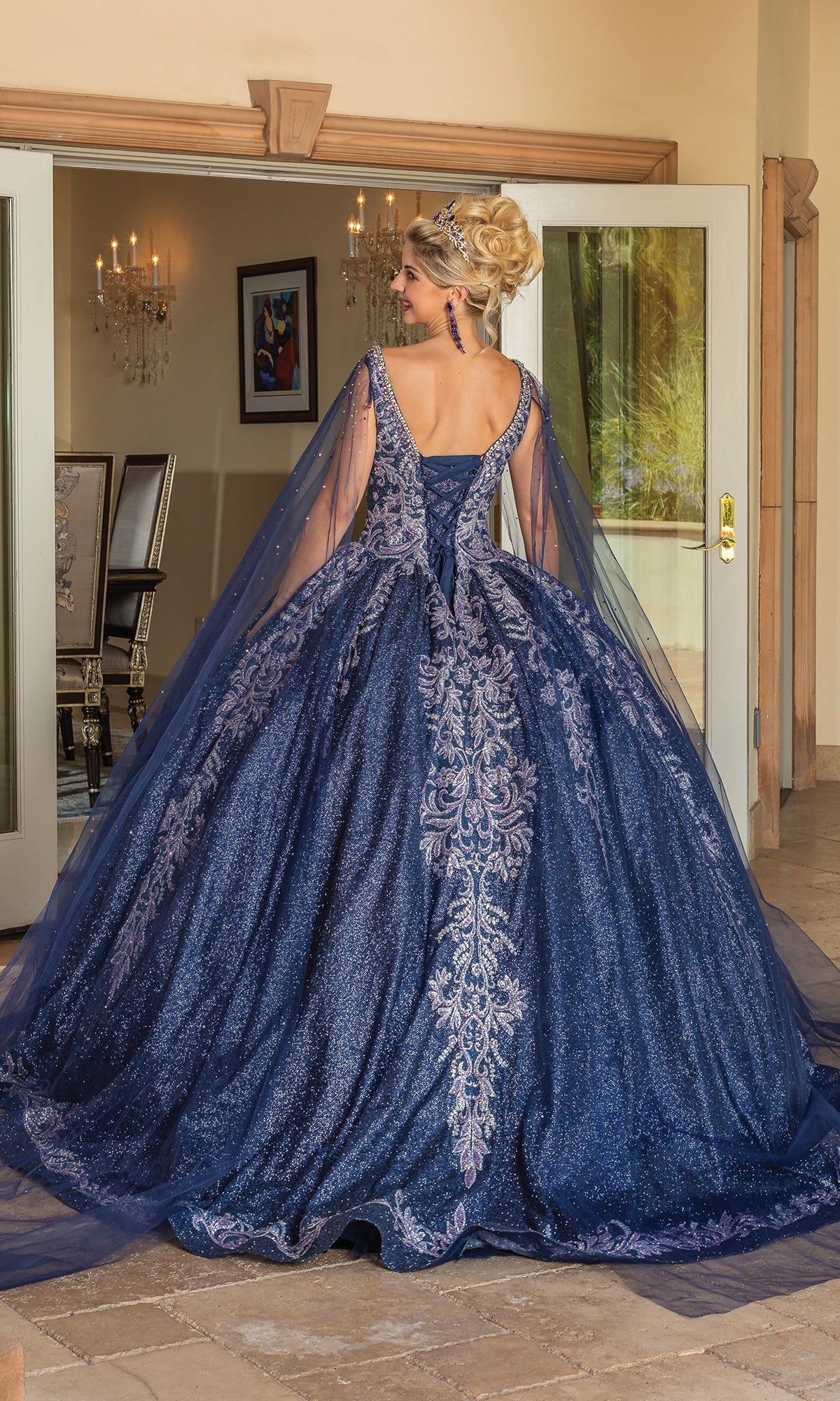 A woman stands indoors facing away, her hair in an elegant updo, wearing the Quinceanera Dress 1751 By Dancing Queen—a navy blue ball gown with intricate lace embroidery and long draped sleeves.