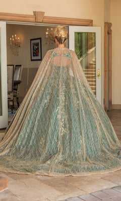 A woman with blonde hair in an updo stands facing away, wearing the Quinceanera Dress 1755 By Dancing Queen, a hunter green gown with gold accents and a sheer embellished cape, inside a building before glass doors.
