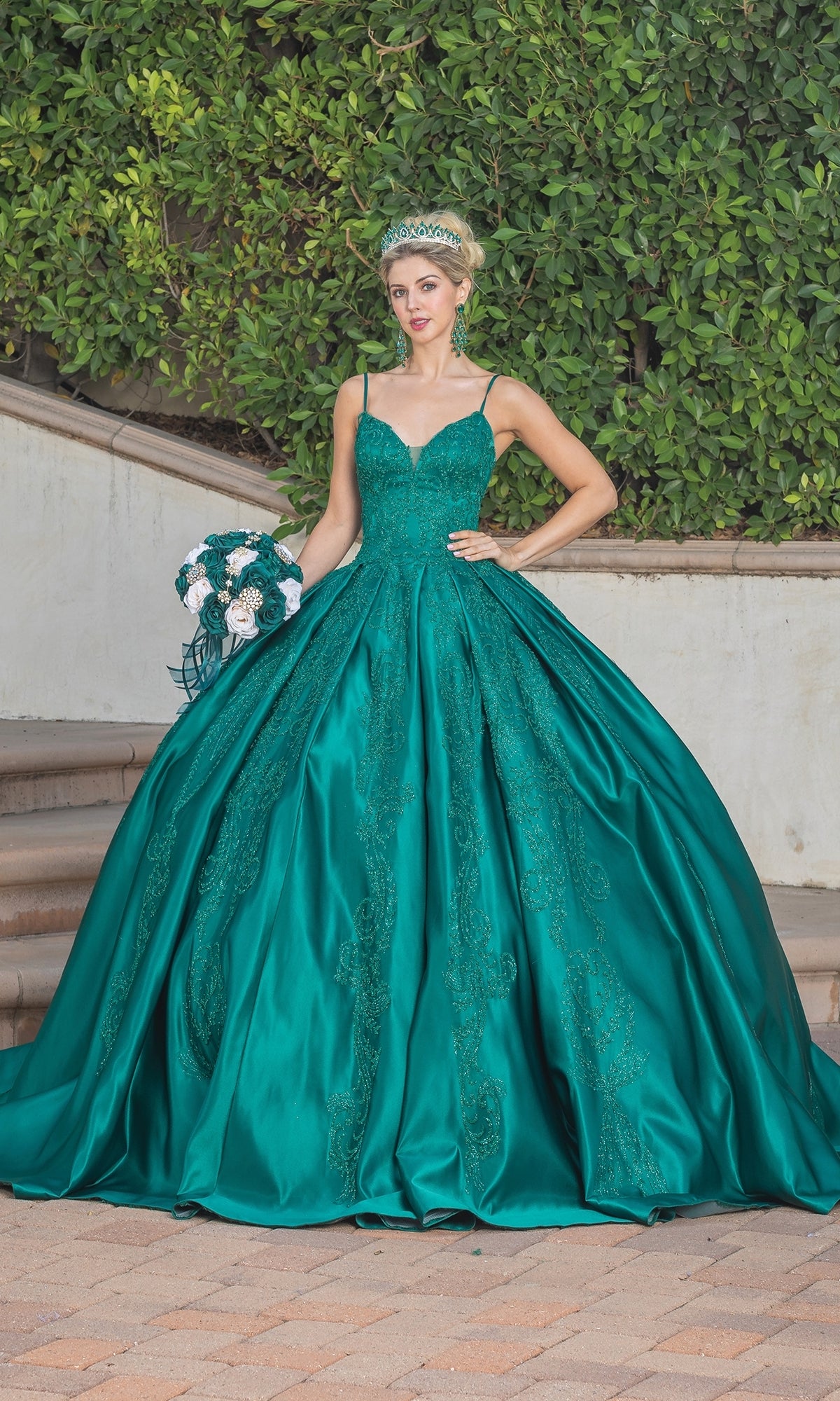 A woman wearing the Quinceanera Dress 1756 By Dancing Queen and a tiara stands outdoors, holding a bouquet of white and teal flowers in front of lush green foliage.