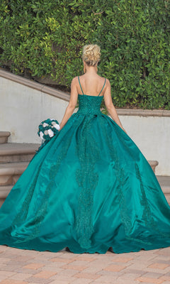 A woman in the Quinceanera Dress 1756 By Dancing Queen, a teal a-line ball gown with lace embroidery, stands on steps holding a bouquet, facing away amid lush green foliage.