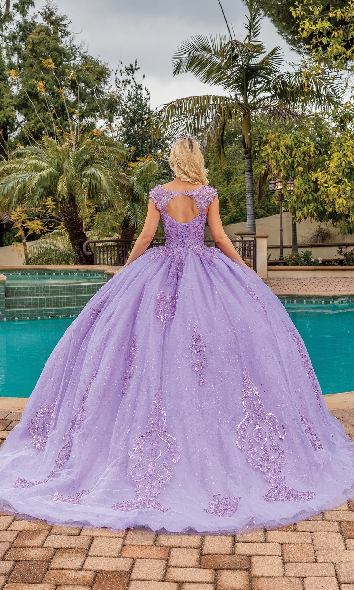 A woman stands by a pool, back to the camera, wearing the Quinceanera Dress 1758 By Dancing Queen—a floor-length lavender a-line ball gown with glitter lace embroidery—surrounded by palm trees and greenery.