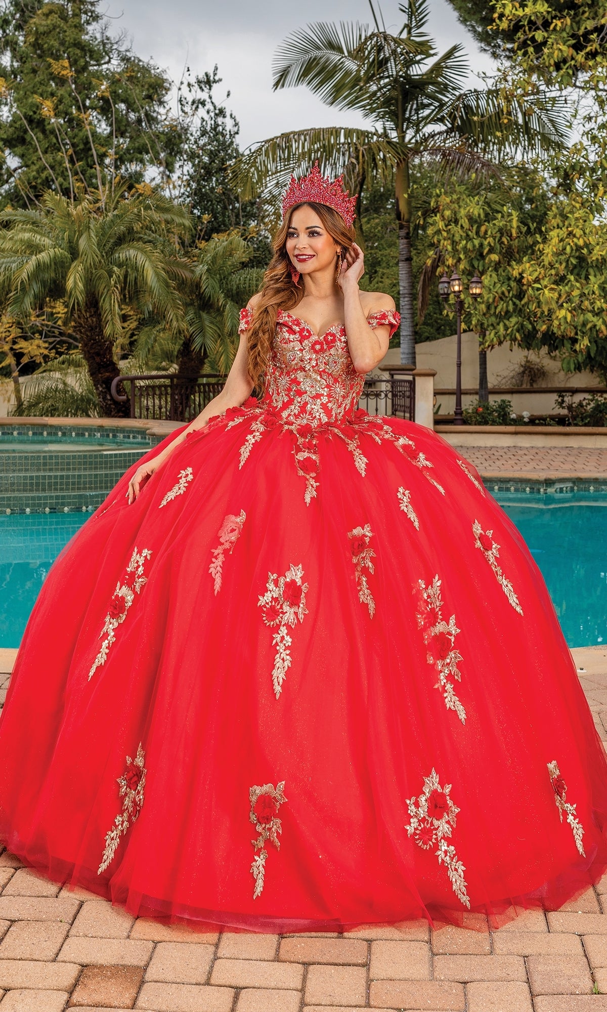 A young woman wears the Quinceanera Dress 1760 By Dancing Queen—a bright red a-line ball gown with gold floral embroidery and a matching crown—while standing by a pool amid lush greenery.
