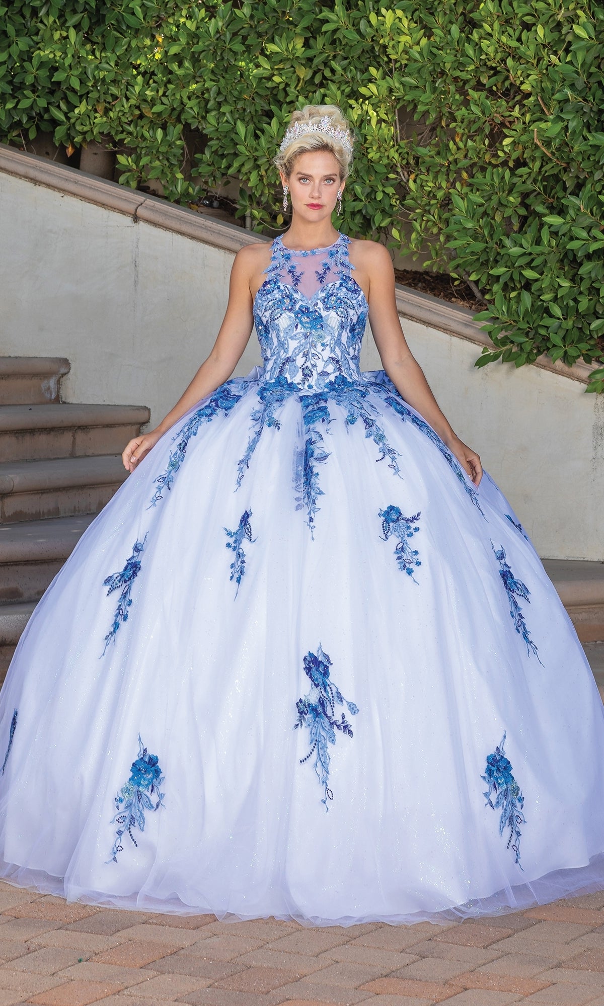 A woman poses outdoors in the Quinceanera Dress 1761 by Dancing Queen, a white ball gown with blue floral and lace embroidery, a fitted bodice, full skirt, bow back detail, and her blonde hair styled up as she gently lifts the skirt.