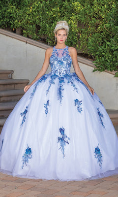 A woman poses outdoors in the Quinceanera Dress 1761 by Dancing Queen, a white ball gown with blue floral and lace embroidery, a fitted bodice, full skirt, bow back detail, and her blonde hair styled up as she gently lifts the skirt.