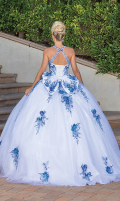 A woman with blonde hair in an updo wears the Quinceanera Dress 1761 By Dancing Queen, featuring blue floral patterns and a bow back detail, while standing outdoors near stairs and greenery.