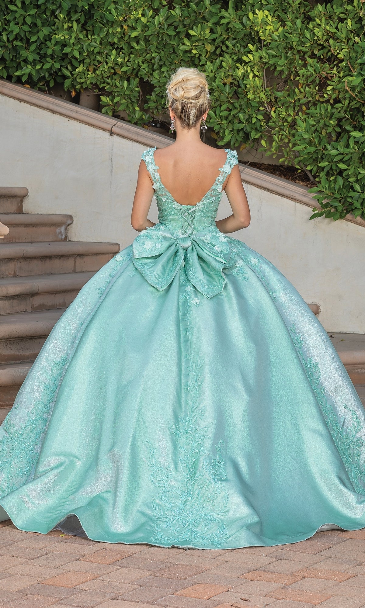 A woman stands outdoors near steps and greenery, facing away, wearing the Dancing Queen Quinceanera Dress 1762—a light blue ball gown with a large back bow and an elegant a-line silhouette.