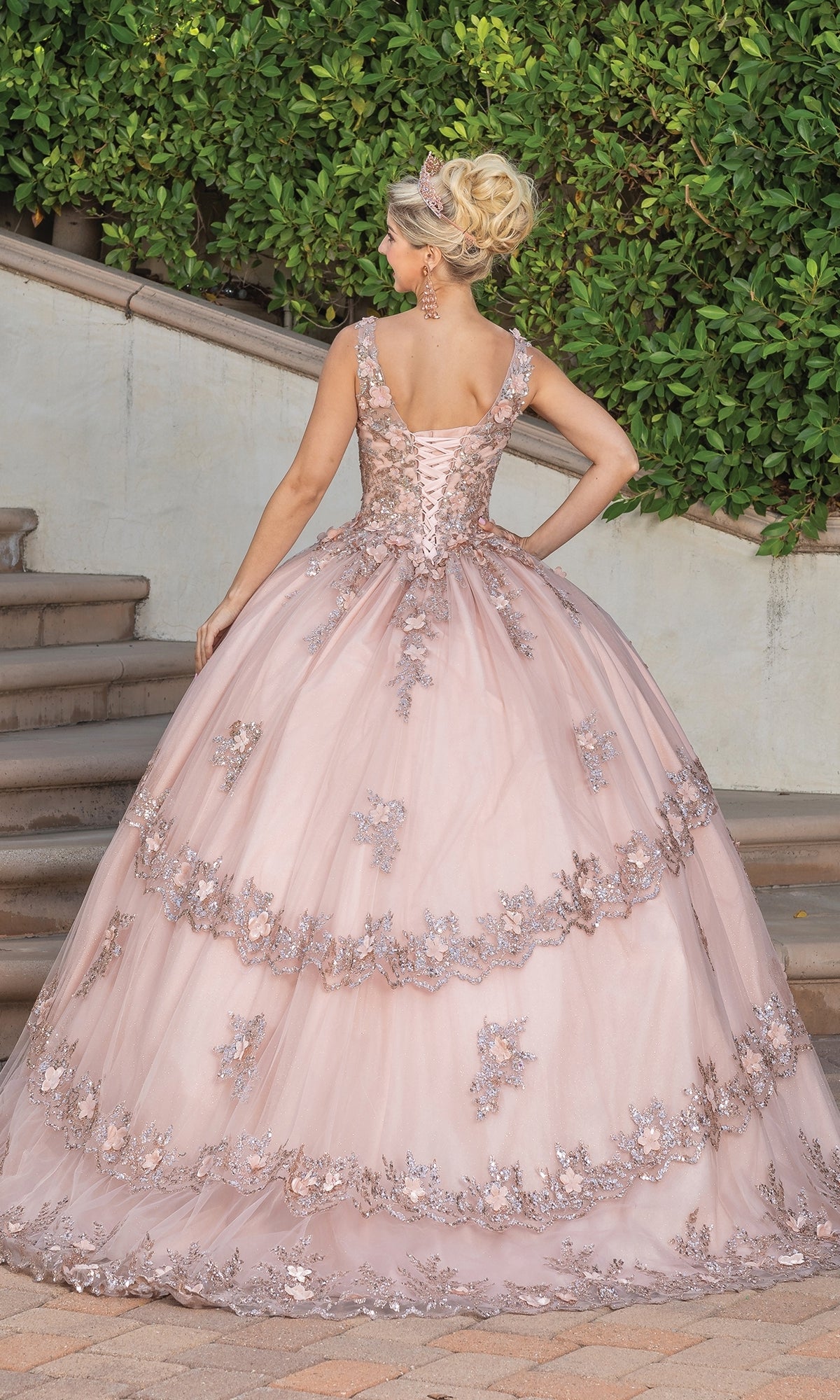 A woman stands on stone steps, facing away, wearing the Quinceanera Dress 1763 By Dancing Queen—a light pink a-line ball gown with floral applique and a voluminous skirt. Green shrubs and a stucco wall complete the charming backdrop.
