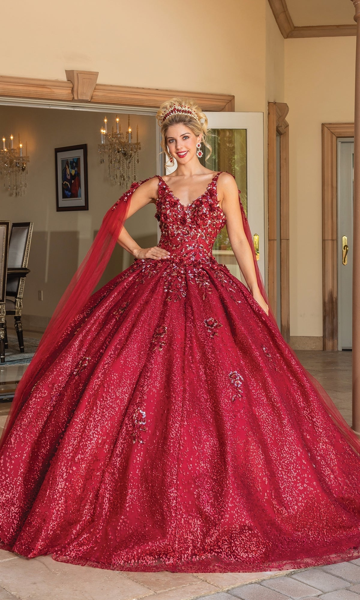 A woman stands indoors wearing the Quinceanera Dress 1764 By Dancing Queen, a sparkling burgundy red ball gown with floral embellishments, v-neckline, tiara, and matching red tulle shoulder accents.