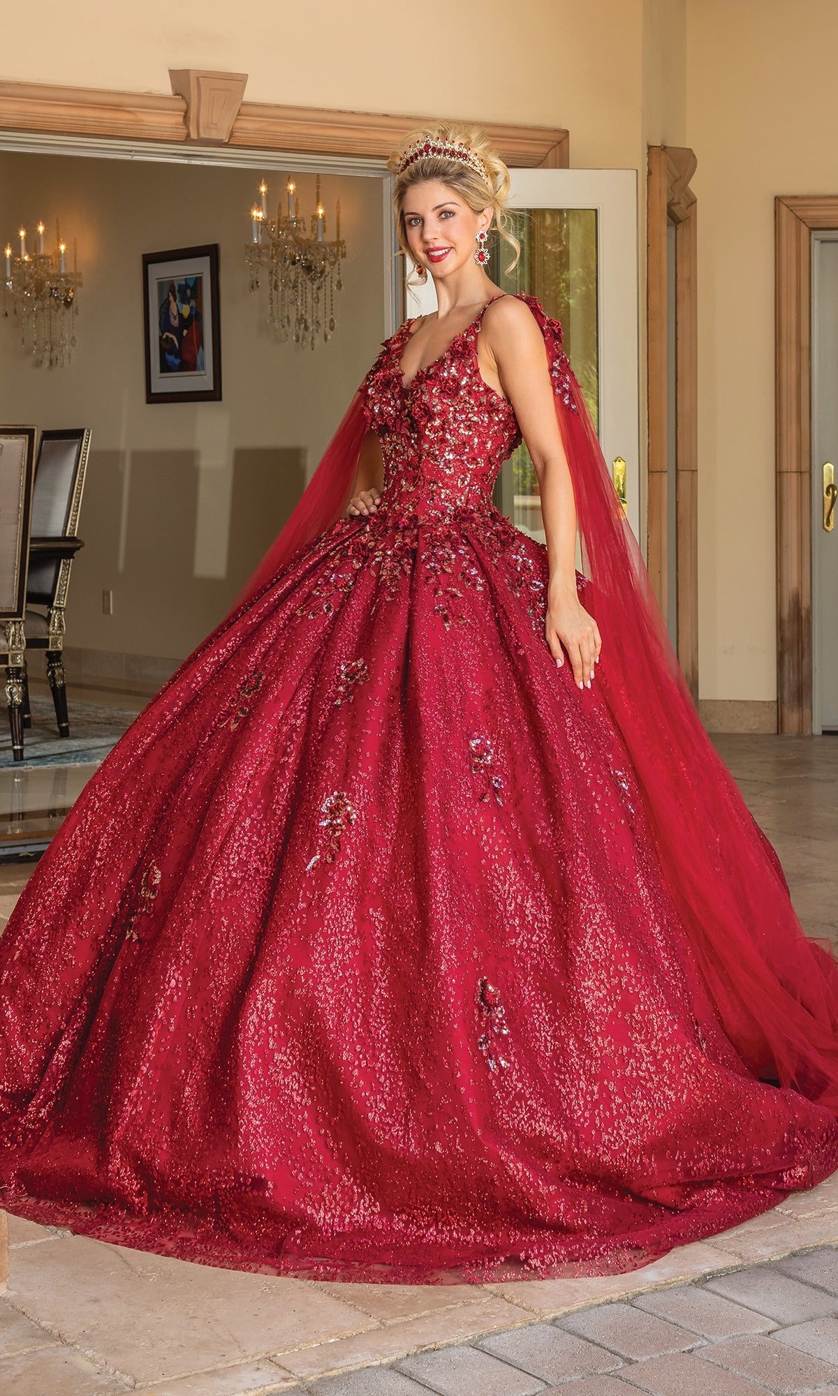 A woman stands indoors smiling at the camera, wearing the Quinceanera Dress 1764 By Dancing Queen, a sparkling burgundy ball gown with floral details and elegant cape sleeves, paired with a tiara.