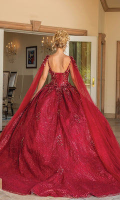 A woman with blonde hair in an updo wears the Quinceanera Dress 1764 By Dancing Queen, a sparkling burgundy ball gown with cape sleeves and a full skirt, standing indoors and facing away from the camera.