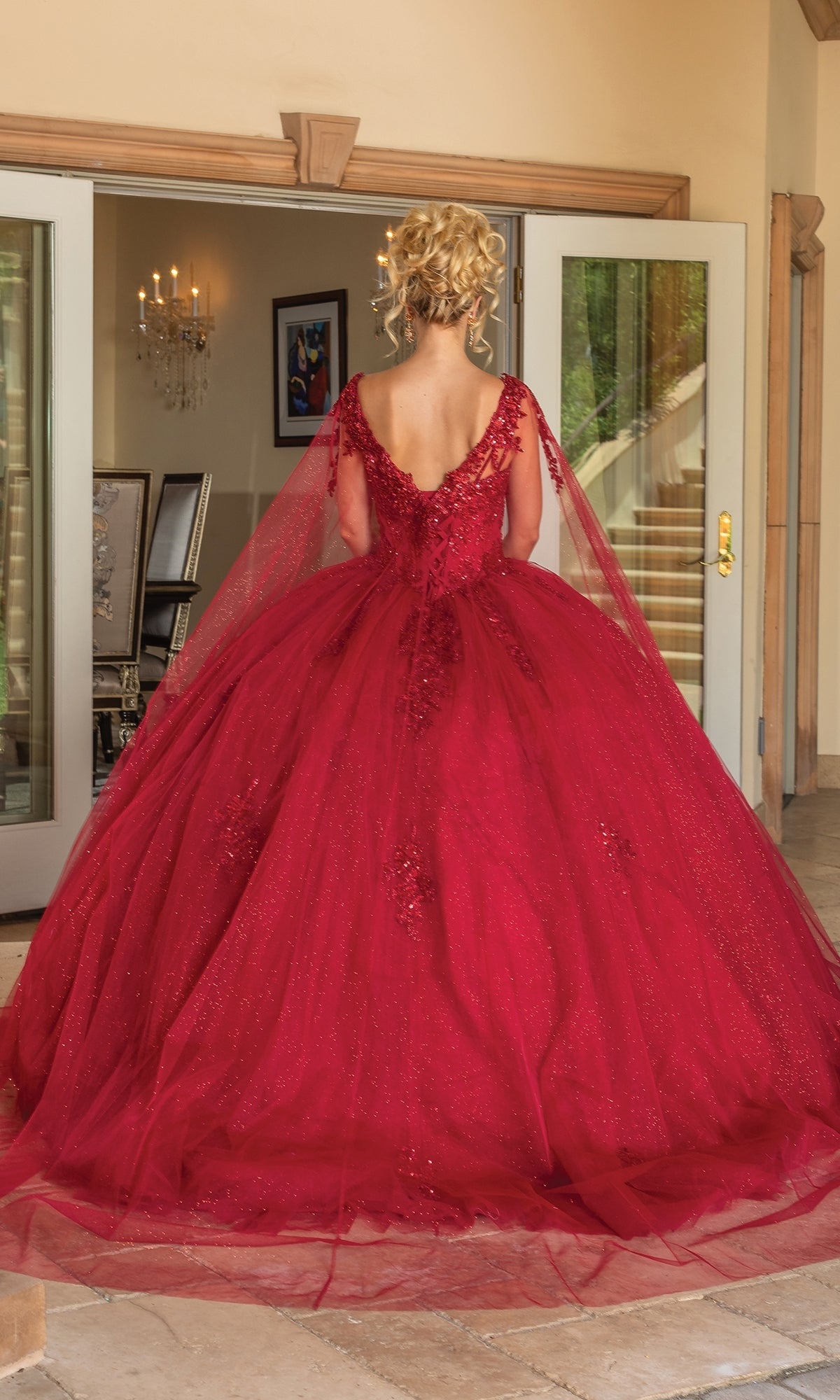 A person stands indoors facing away, wearing the Quinceanera Dress 1767 By Dancing Queen—a sparkly red a-line ball gown with a voluminous skirt, deep V-back, and sheer tulle overlay. Double doors and a chandelier are in the background.