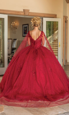 A person stands indoors facing away, wearing the Quinceanera Dress 1767 By Dancing Queen—a sparkly red a-line ball gown with a voluminous skirt, deep V-back, and sheer tulle overlay. Double doors and a chandelier are in the background.