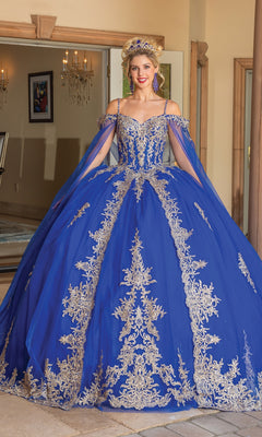 A woman poses gracefully indoors in the Quinceanera Dress 1768 By Dancing Queen—a royal blue off-the-shoulder ball gown with gold embroidery, matching accessories, and a tiara—standing near an open doorway.