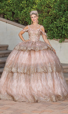 A woman stands outdoors in the Quinceanera Dress 1770 by Dancing Queen, a glittery pink and gold A-line ball gown with layered ruffles, off-shoulder straps, and a sweetheart neckline, accessorized with a tiara and earrings.