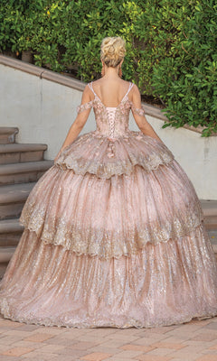 A person in the Quinceanera Dress 1770 By Dancing Queen, a pink and gold glittery A-line ball gown with tiered layers, stands outdoors near steps and greenery, facing away from the camera.