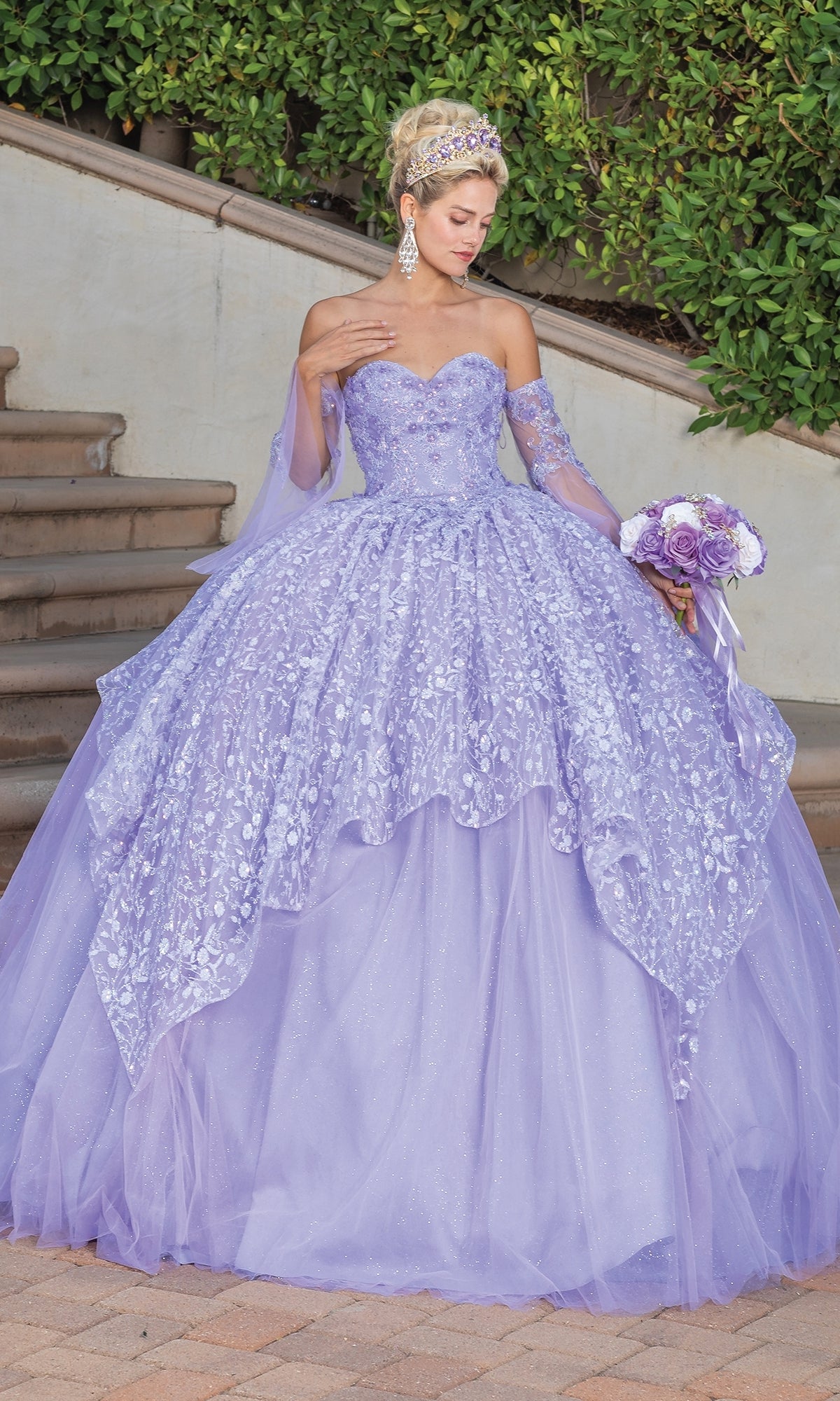 A woman in the Quinceanera Dress 1773 by Dancing Queen, a strapless ball gown with floral patterns, stands on a path holding a matching bouquet, with steps and greenery in the background.