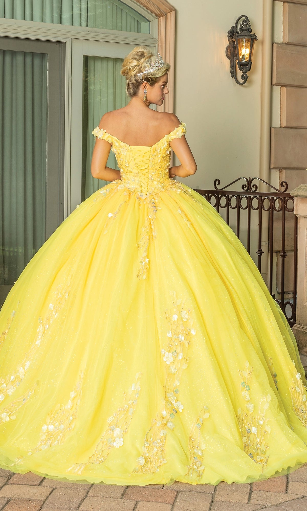 Outdoors by a window, a woman wears the Quinceanera Dress 1775 By Dancing Queen—a yellow ball gown featuring floral details, lace embroidery, off-the-shoulder sleeves, and a voluminous skirt.