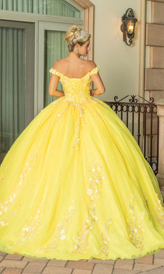 Outdoors by a window, a woman wears the Quinceanera Dress 1775 By Dancing Queen—a yellow ball gown featuring floral details, lace embroidery, off-the-shoulder sleeves, and a voluminous skirt.