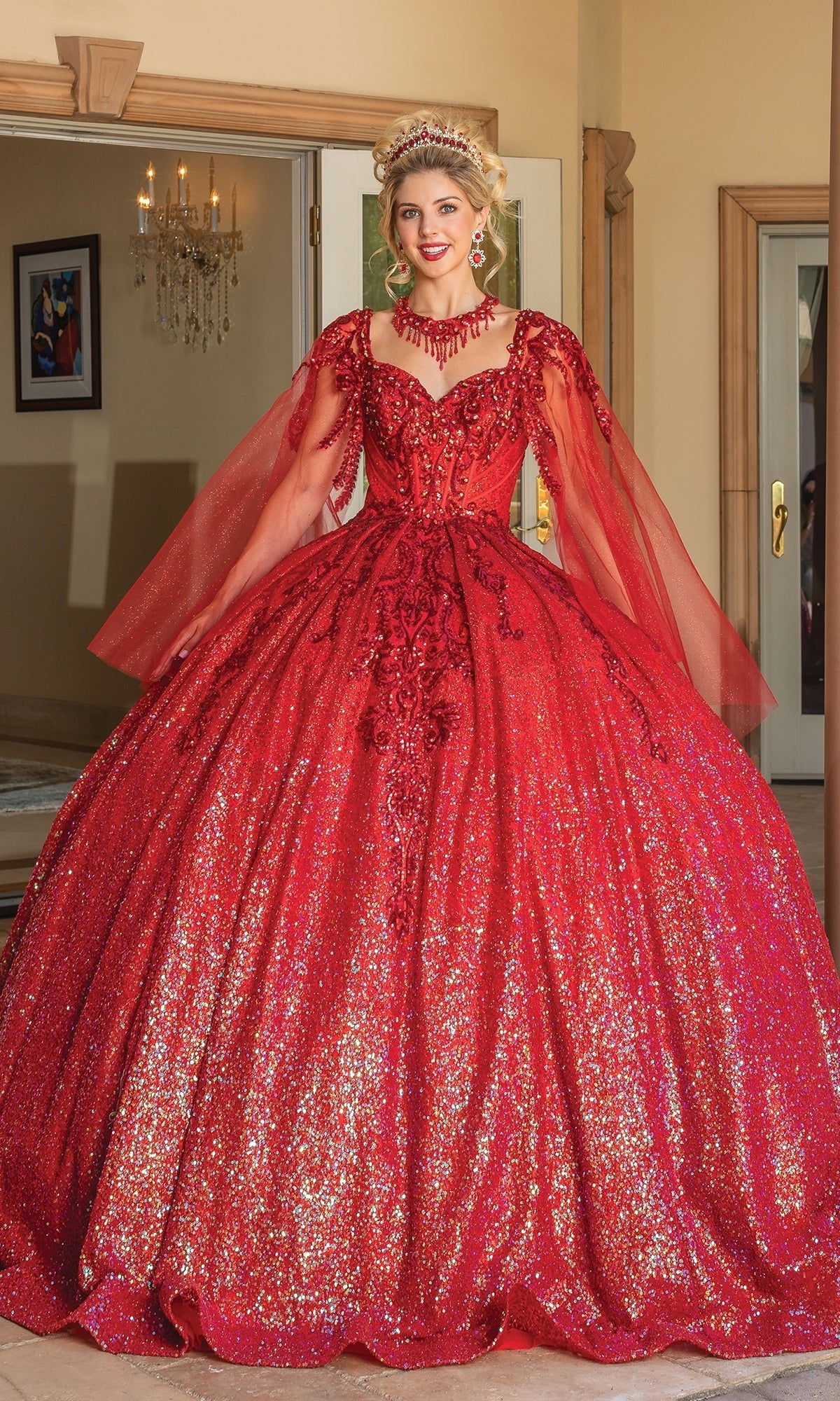 A woman stands indoors wearing the Quinceanera Dress 1776 By Dancing Queen, a sparkling red a-line gown with sheer long sleeves, detailed embroidery, and matching red jewelry.