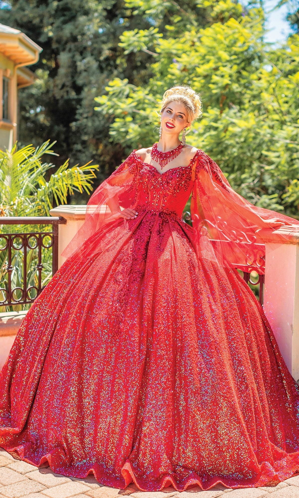 A woman stands outdoors in the Quinceanera Dress 1776 By Dancing Queen, a sparkling red A-line gown with sheer long sleeves and intricate embellishments, surrounded by greenery and sunlight.
