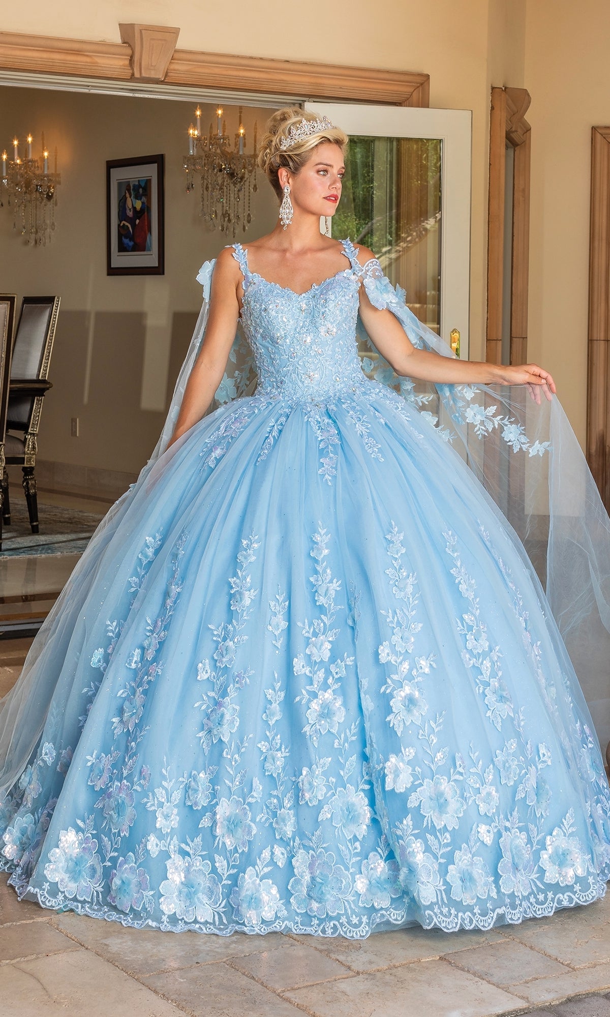 A woman stands indoors holding the edge of her Quinceanera Dress 1778 By Dancing Queen—a light blue ball gown with a sweetheart neckline, floral embroidery, and a full skirt.