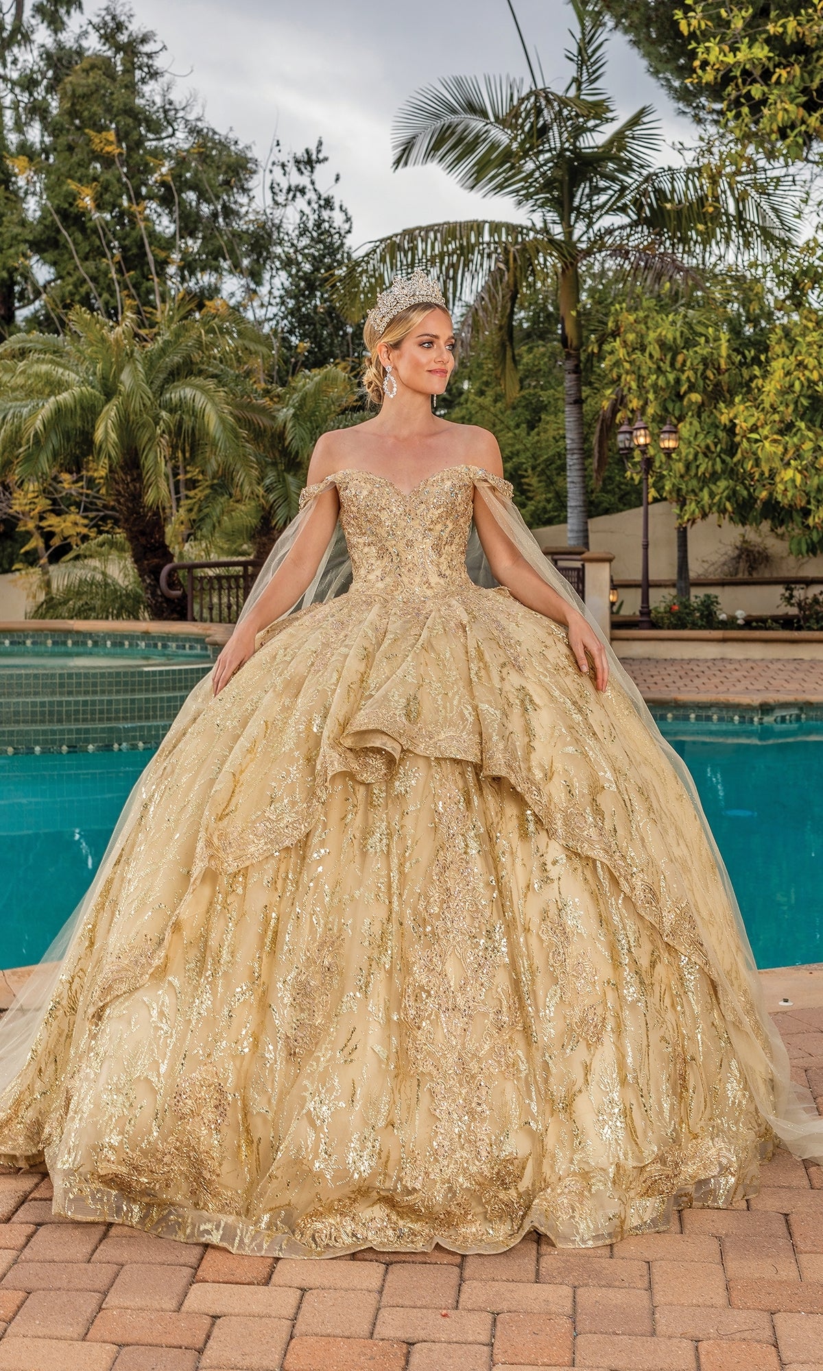 A woman poses by a pool among palm trees wearing Quinceanera Dress 1782 By Dancing Queen, an elaborate gold off-the-shoulder gown with a sweetheart neckline and gold crown.