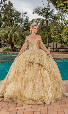 A woman poses by a pool among palm trees wearing Quinceanera Dress 1782 By Dancing Queen, an elaborate gold off-the-shoulder gown with a sweetheart neckline and gold crown.