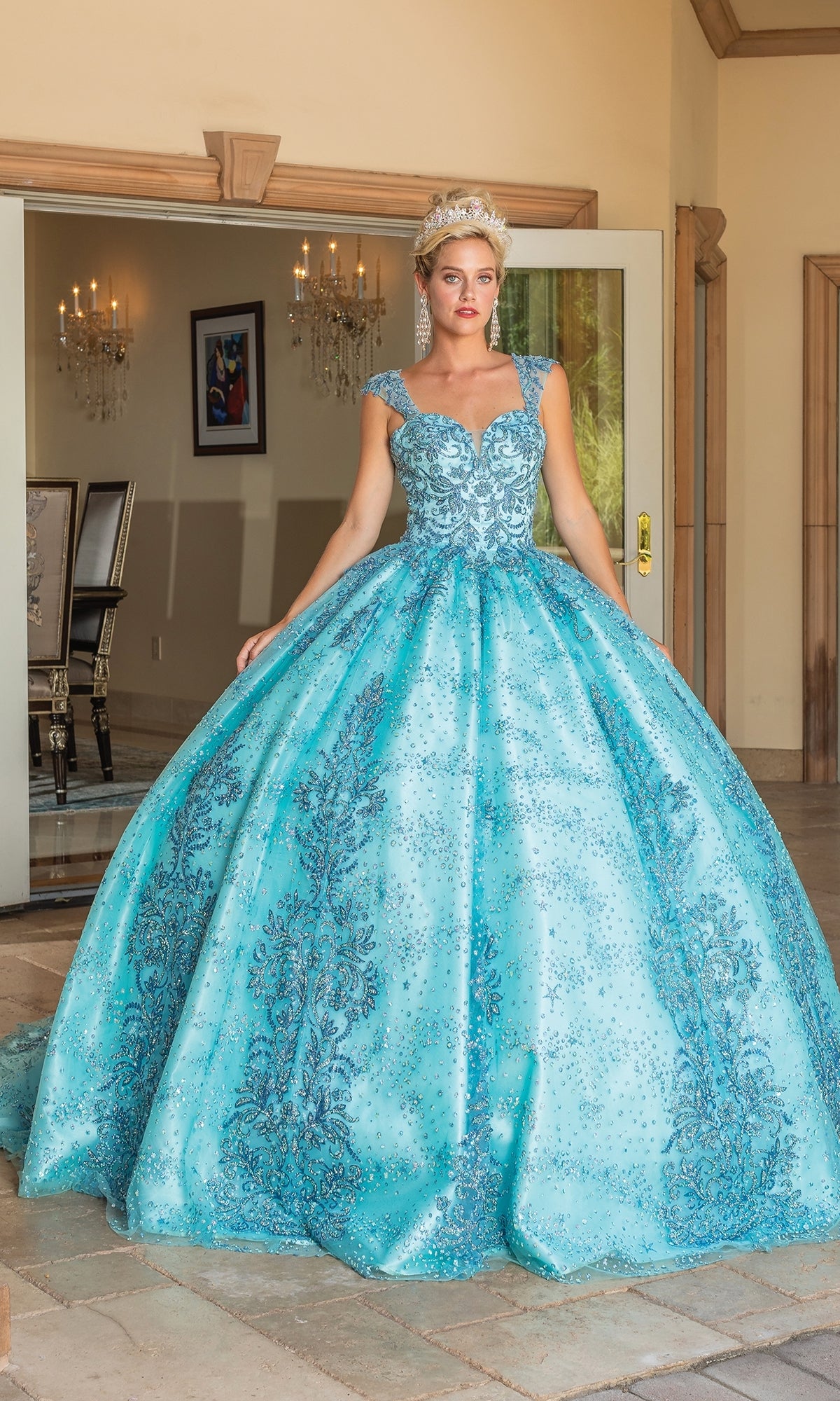 A woman stands in an elegant room wearing Quinceanera Dress 1784 By Dancing Queen, a sparkling aqua blue ball gown with silver embroidery and a sweetheart neckline, accessorized with a tiara.