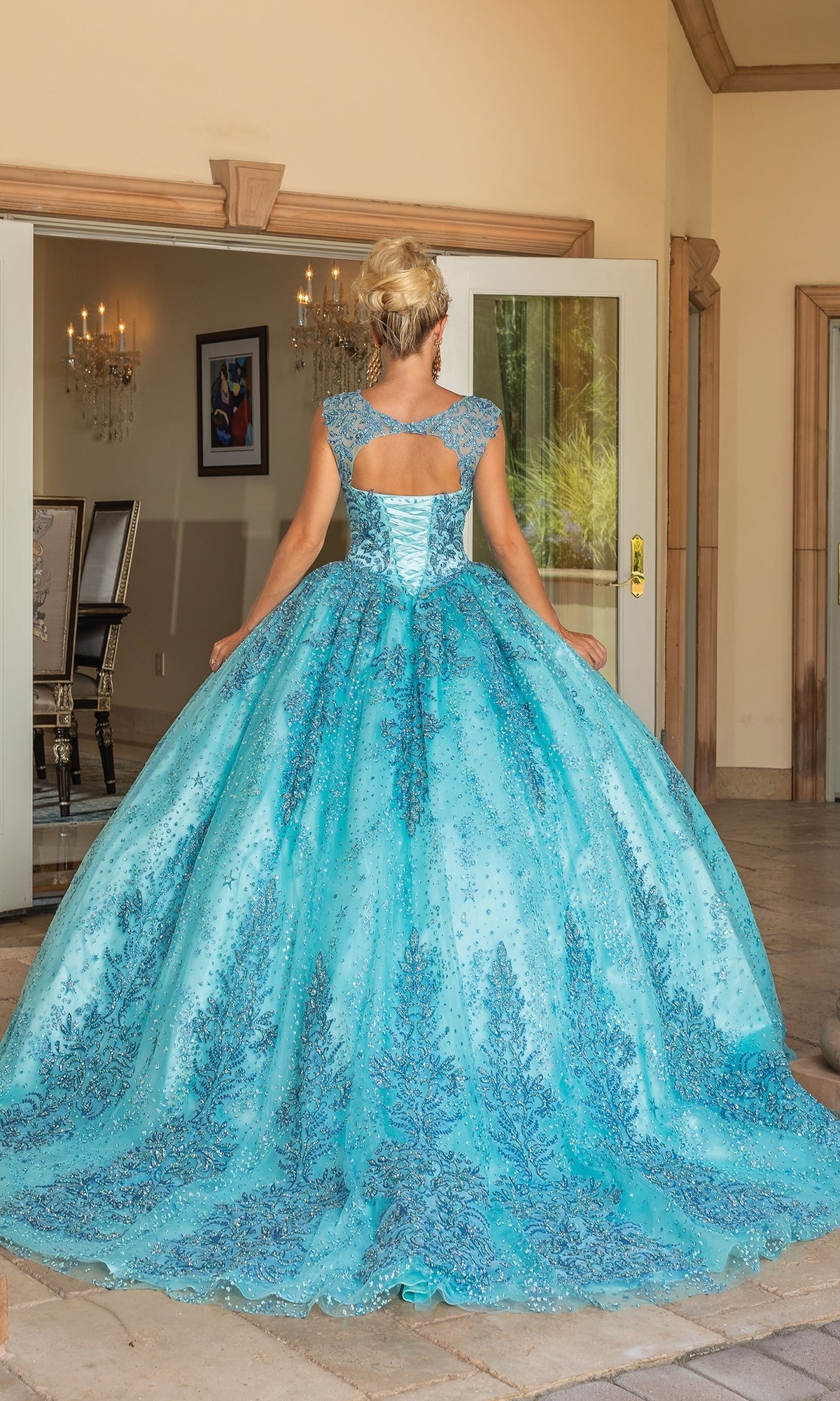 A woman stands facing away in the Dancing Queen Quinceanera Dress 1784, a voluminous blue ball gown with intricate silver accents and a sweetheart neckline, inside an elegant room with an open doorway.