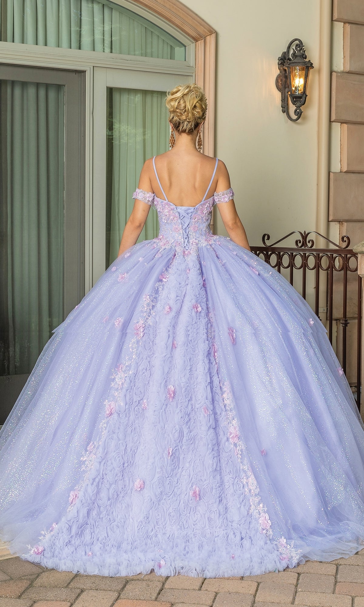 A person stands outdoors facing away, wearing the Quinceanera Dress 1785 By Dancing Queen—a lilac ball gown with floral details and off-the-shoulder straps—hair elegantly styled in an updo.