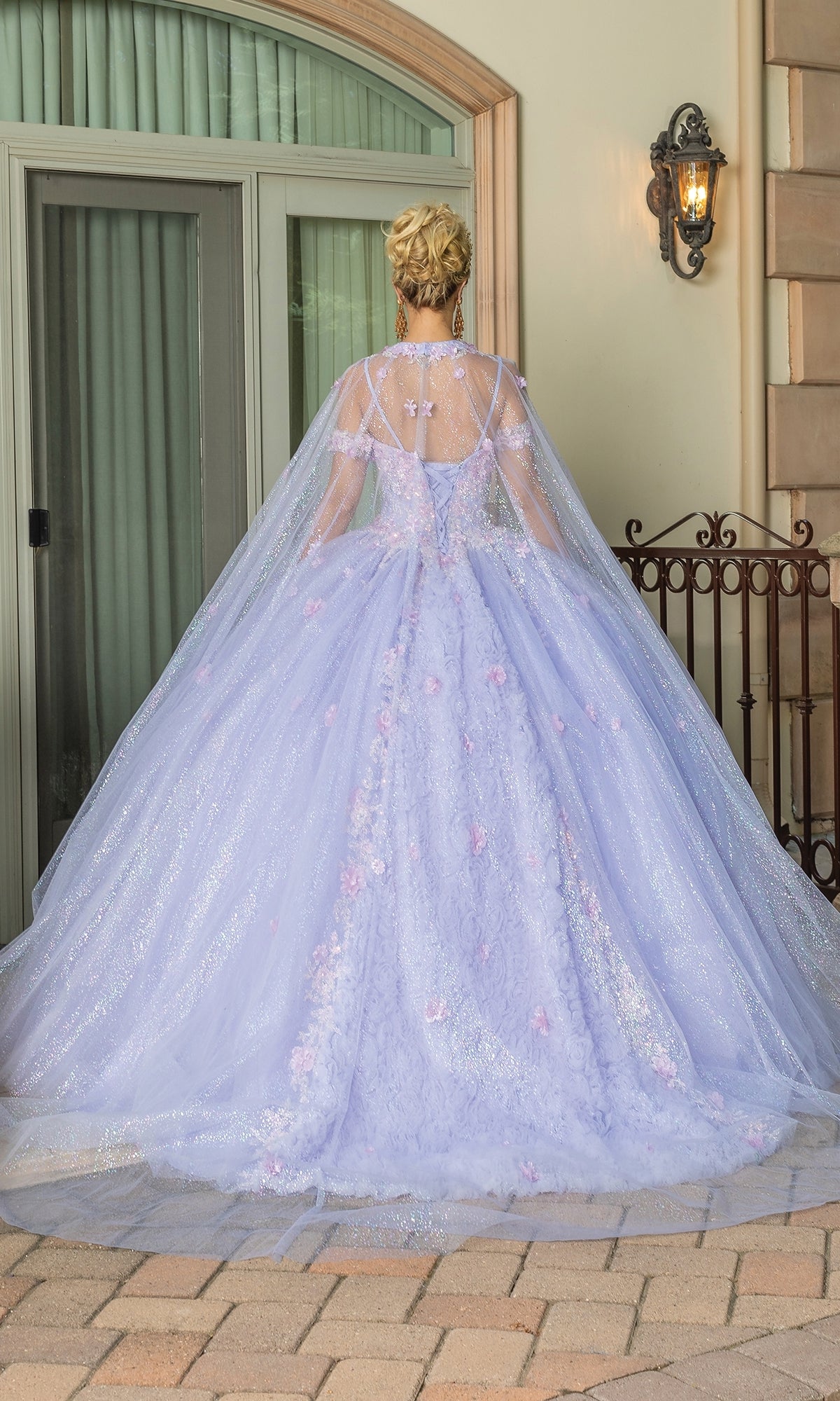 A woman stands outside facing away, wearing the Quinceanera Dress 1785 By Dancing Queen—a lilac ball gown with floral details, a sheer cape, and a sweetheart neckline—by a glass door.