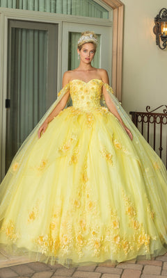 A woman stands by a window and railing wearing Quinceanera Dress 1788 By Dancing Queen, a yellow off-the-shoulder floral A-line ball gown with a sweetheart neckline and sheer cape.