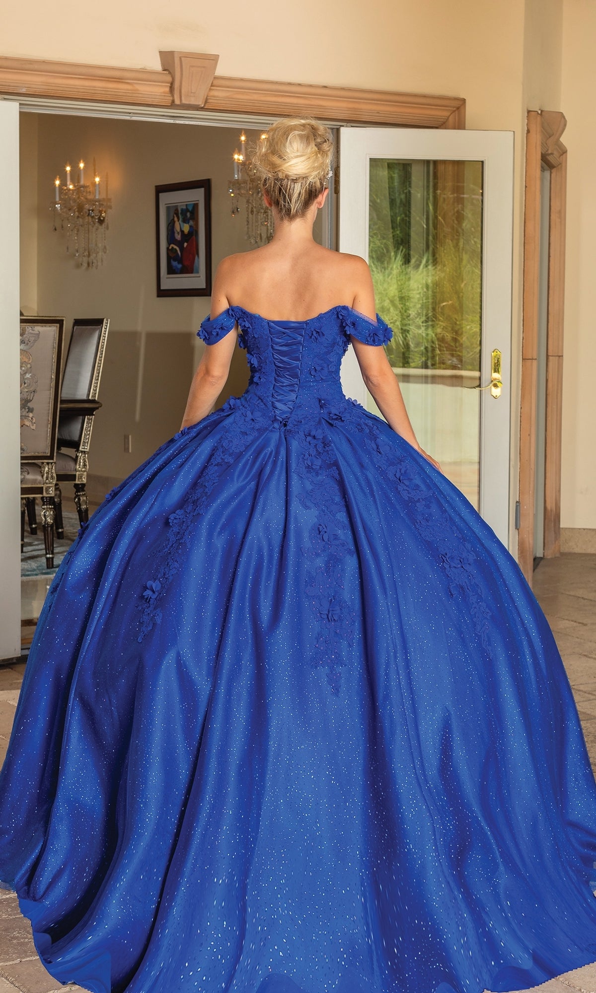 A woman stands indoors facing away, wearing the Quinceanera Dress 1790 By Dancing Queen—a royal blue gown with an off-the-shoulder neckline and full skirt.