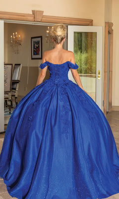 A woman stands indoors facing away, wearing the Quinceanera Dress 1790 By Dancing Queen—a royal blue gown with an off-the-shoulder neckline and full skirt.
