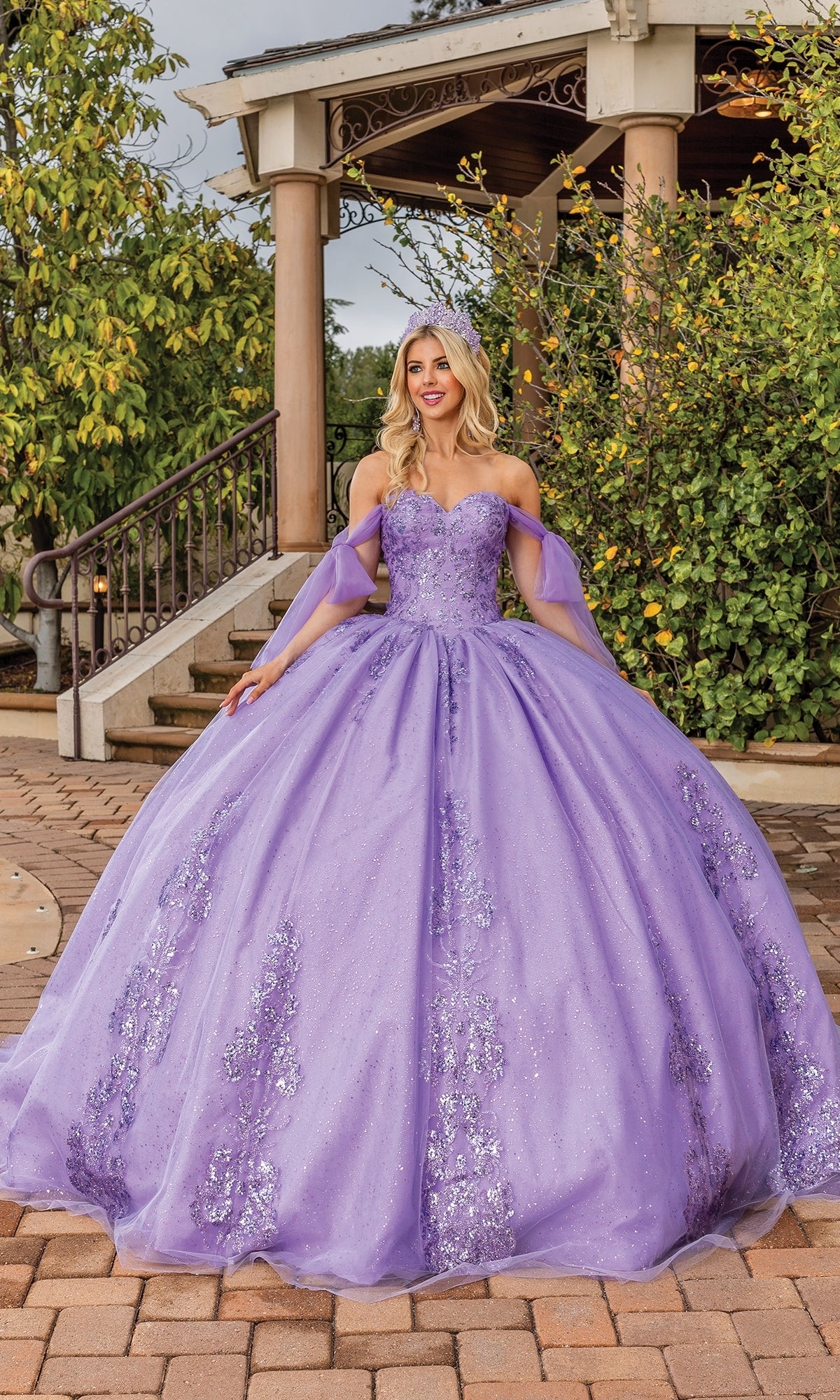A woman stands outdoors in front of greenery and a gazebo, wearing the Quinceanera Dress 1791 By Dancing Queen—an off-the-shoulder lavender ball gown with silver embellishments and elegant sleeves.