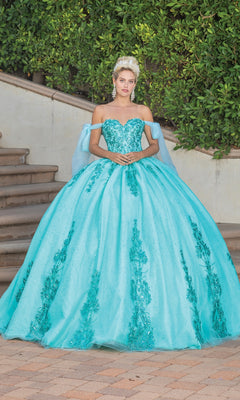 Outdoors before stairs and greenery, a person wears the Quinceanera Dress 1791 By Dancing Queen—an off-shoulder blue ball gown with sequined patterns—and a tiara.