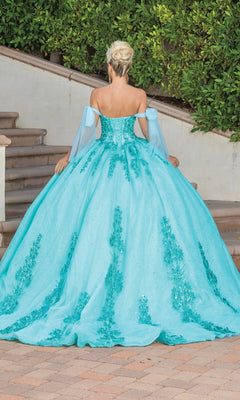 A woman with blonde hair in an updo models the Quinceanera Dress 1791 By Dancing Queen, an off-the-shoulder turquoise ball gown with floral details, standing outside on a tiled walkway near stairs and lush greenery.