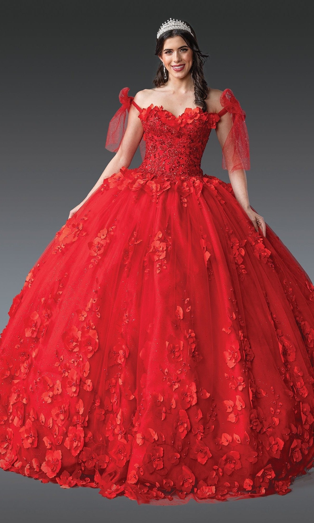 A woman stands gracefully against a plain, dark background, wearing the Quinceanera Dress 1792 by Dancing Queen—a bright red, off-the-shoulder tulle ballgown adorned with beaded floral appliqués and layered tulle—complete with a sparkling tiara.