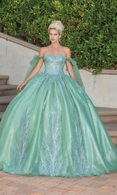 A woman stands outdoors in the Quinceanera Dress 1793 By Dancing Queen, an off-the-shoulder sage green ball gown with silver leaf patterns and a tiara, set amidst lush greenery and stone steps.